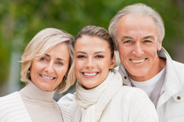 A senior couple and a young woman are in a park setting, smiling towards the camera with their heads together. Horizontal shot.